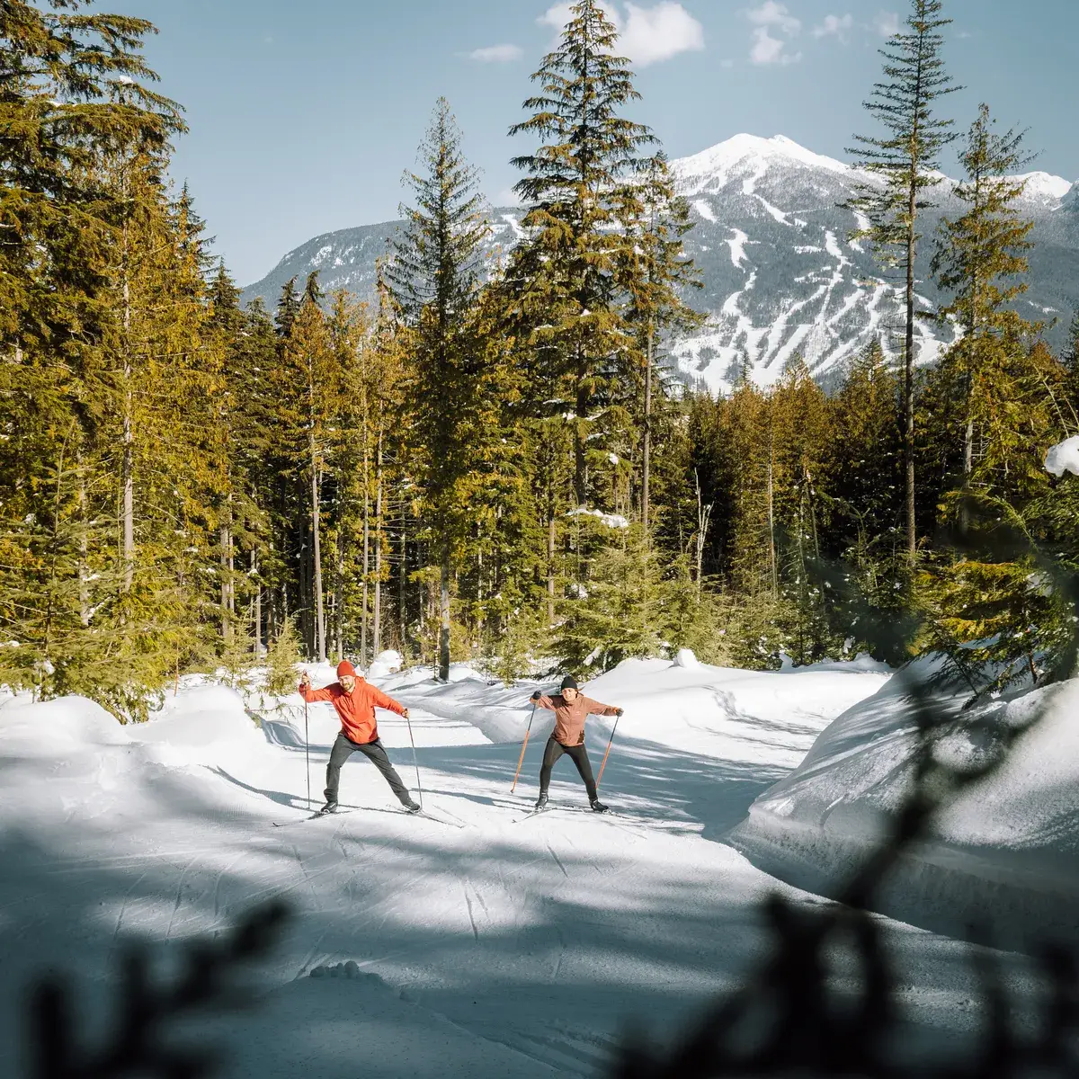 Two people cross-country skiing on a groomed snowy trail surrounded by pine trees with mountains in the background.