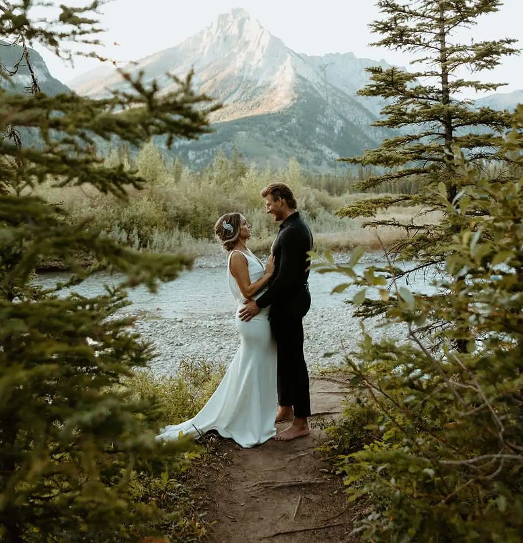 Couple in wedding attire embracing by a river with mountains