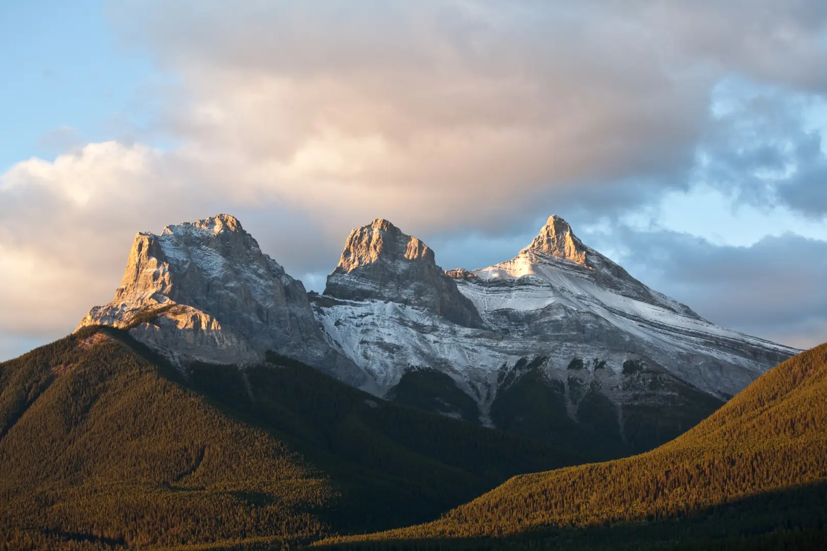 Three Sisters peaks in Canmore, Alberta, with golden hour light on snow-capped summits and pine forests.