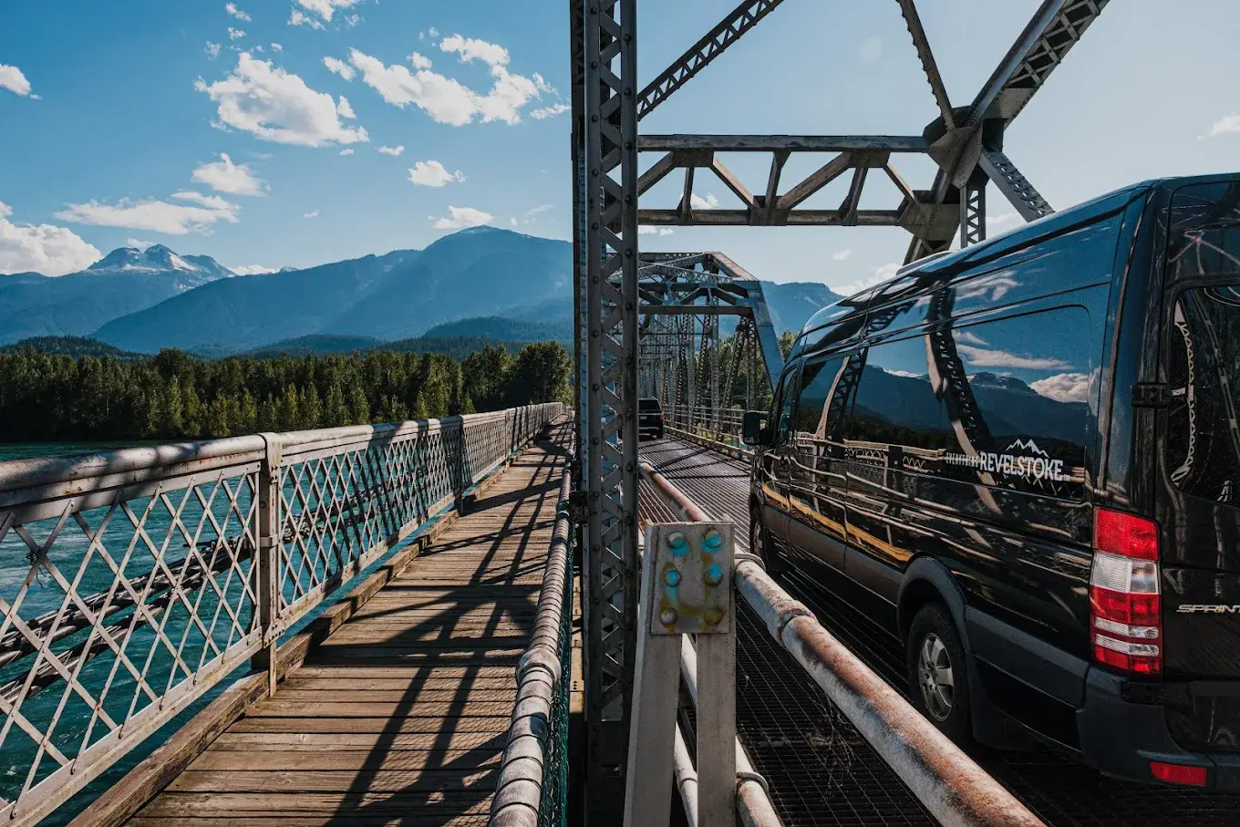 Black Revelstoke van crossing a bridge over a river with mountains in background