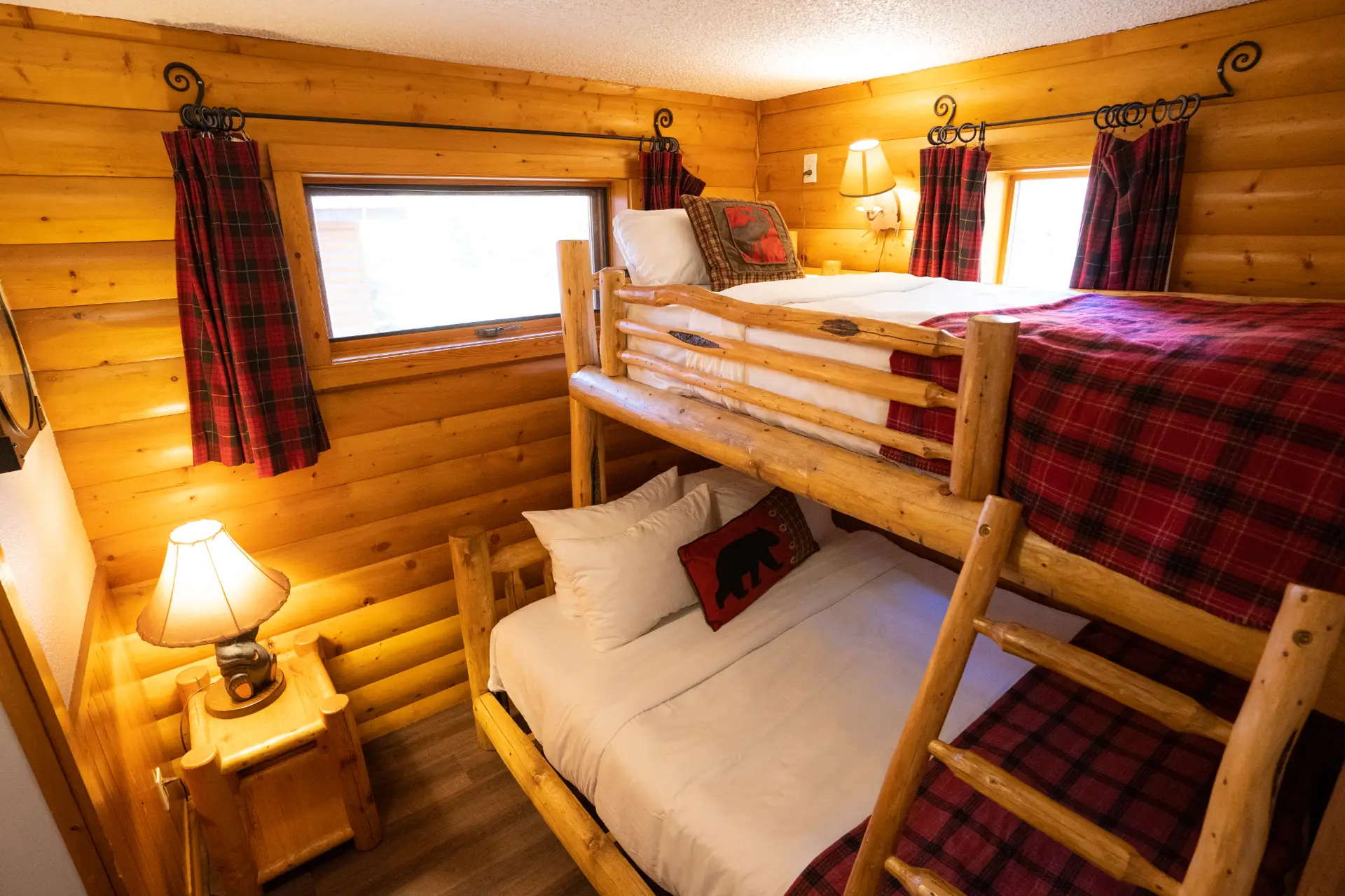 Interior of a rustic log cabin bedroom with bunk beds, plaid blankets, and warm lighting.