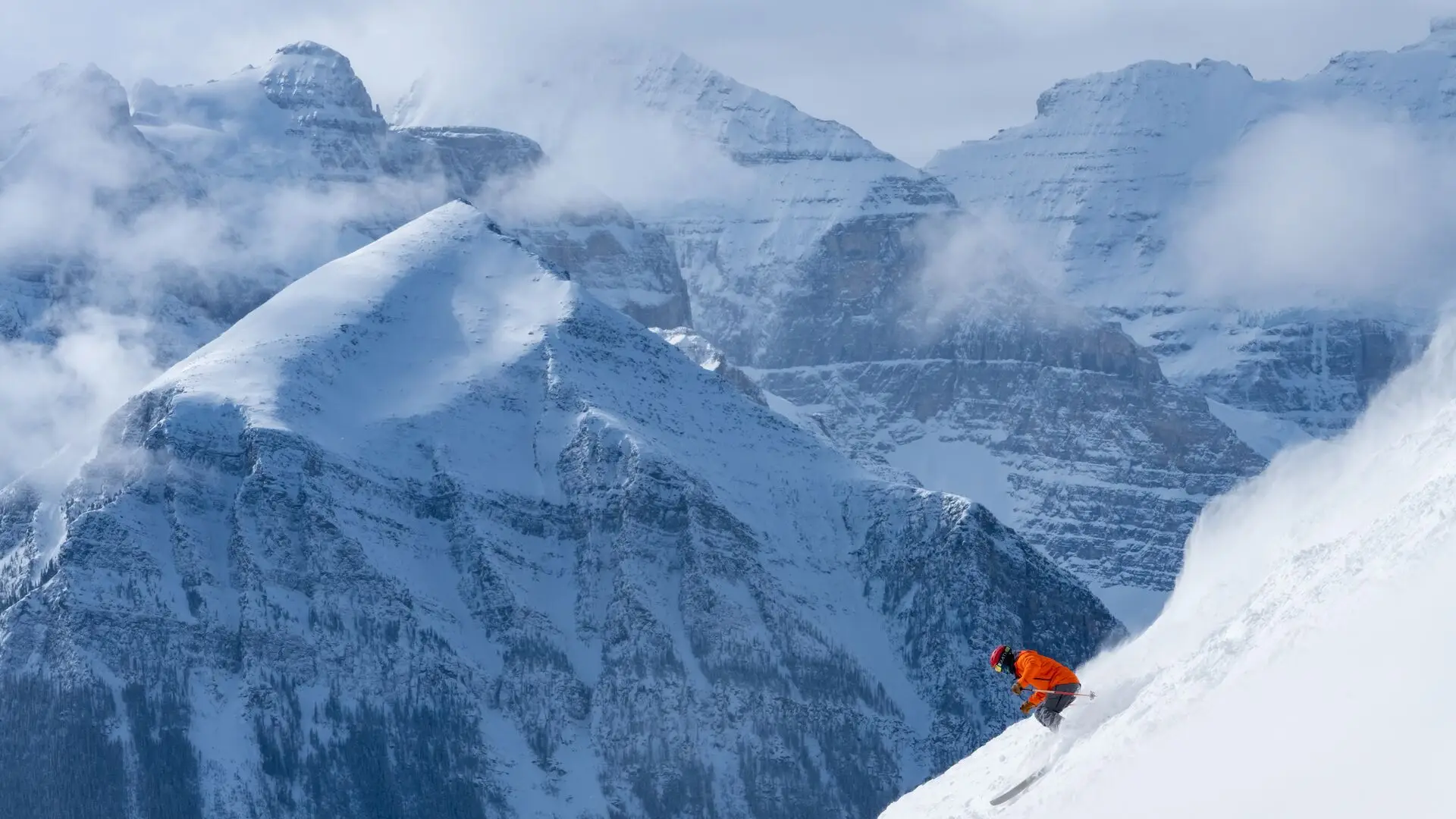 SkiBig3: A skier in an orange jacket carves down a steep, snowy slope with majestic mountains behind.