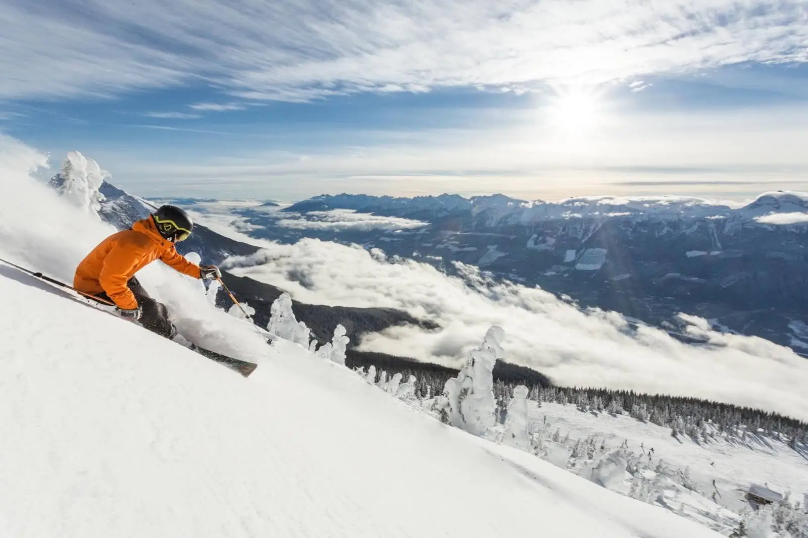 Skier in orange jacket descends snowy slope with mountain and cloud backdrop.